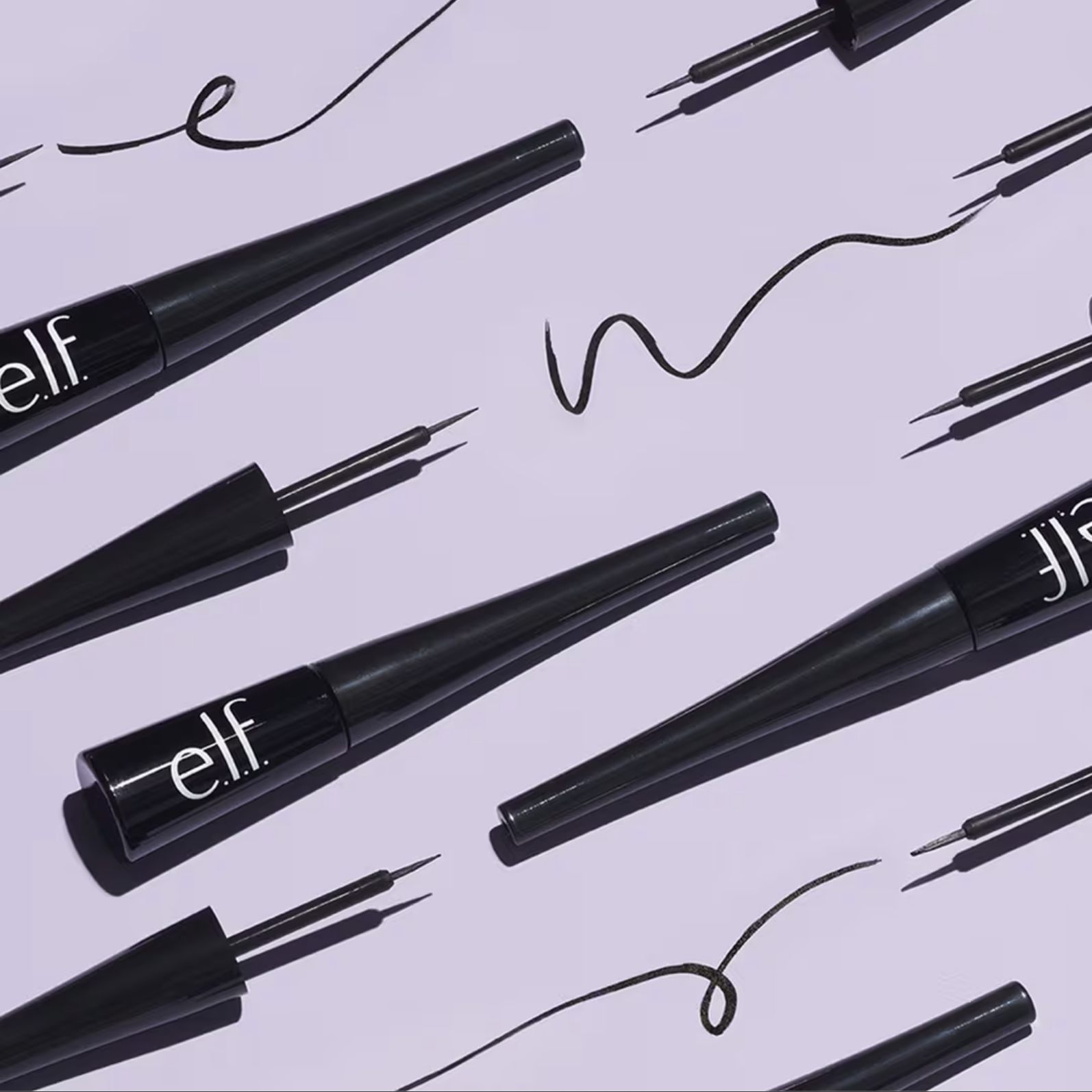 Flat lay of e.l.f. Expert Liquid Eyeliner pens on a lavender background, showing their thin brush tips and black eyeliner strokes